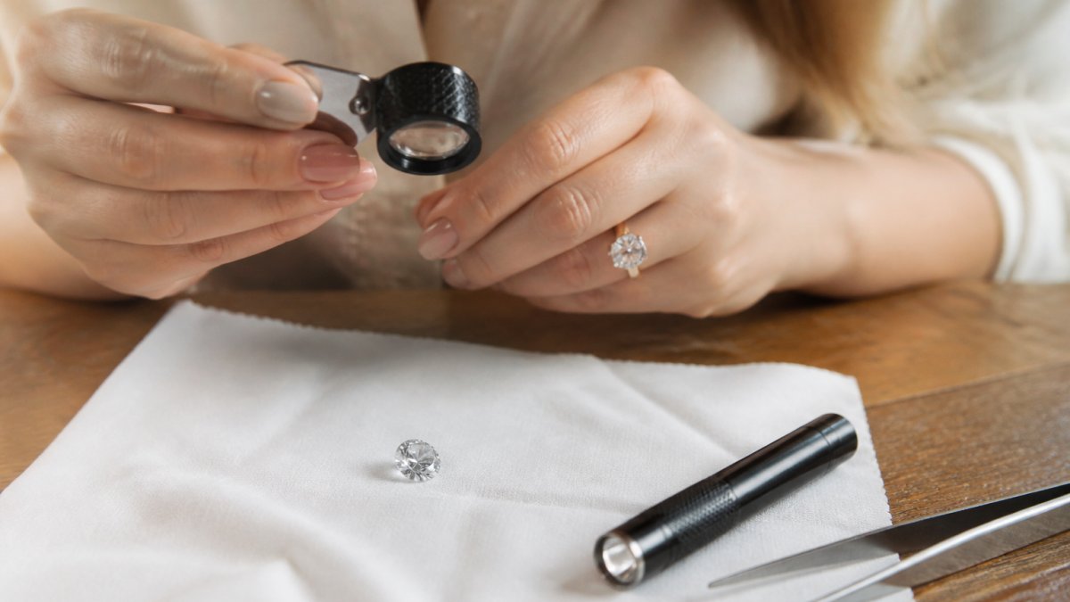 Close-up of a woman's hands holding a jeweler's loupe over a loose moissanite stone on a soft white cloth, with a small penlight and tweezers arranged nearby on a wooden table in warm natural light