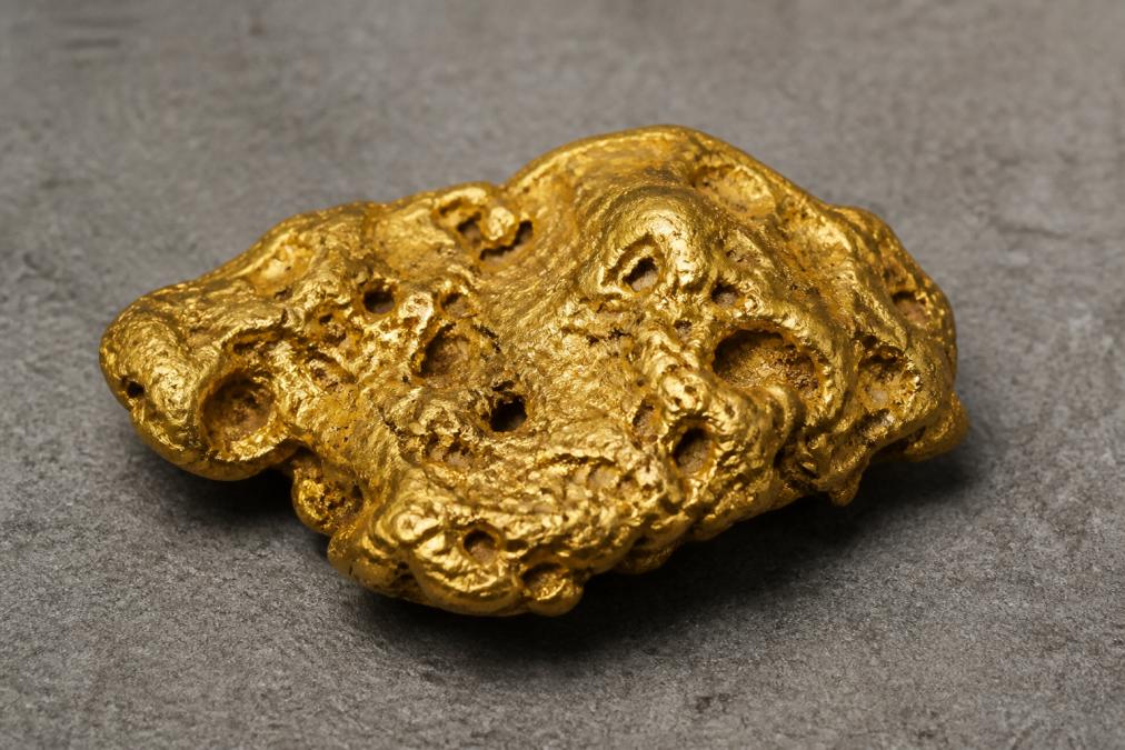 Close-up of natural Australian gold nugget resting on neutral grey surface, showing rich golden metallic luster, uneven surface texture with natural cavities and dimples formed by geological weathering, photographed under warm natural lighting