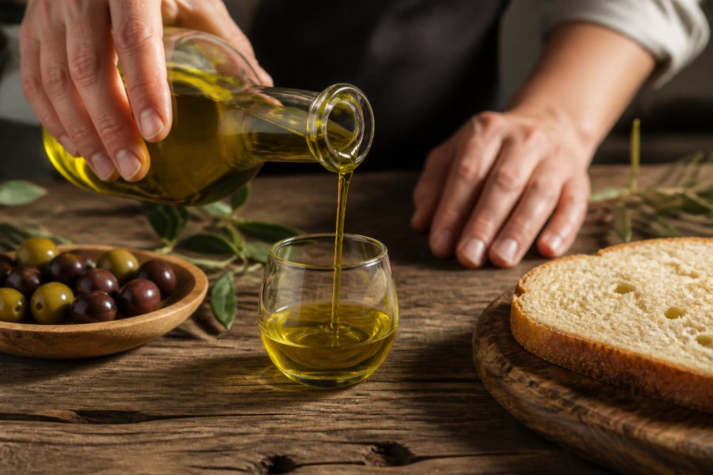 Close-up of hands pouring golden-green olive oil into a small tasting glass on a rustic wooden table, with a slice of bread and a bowl of olives nearby
