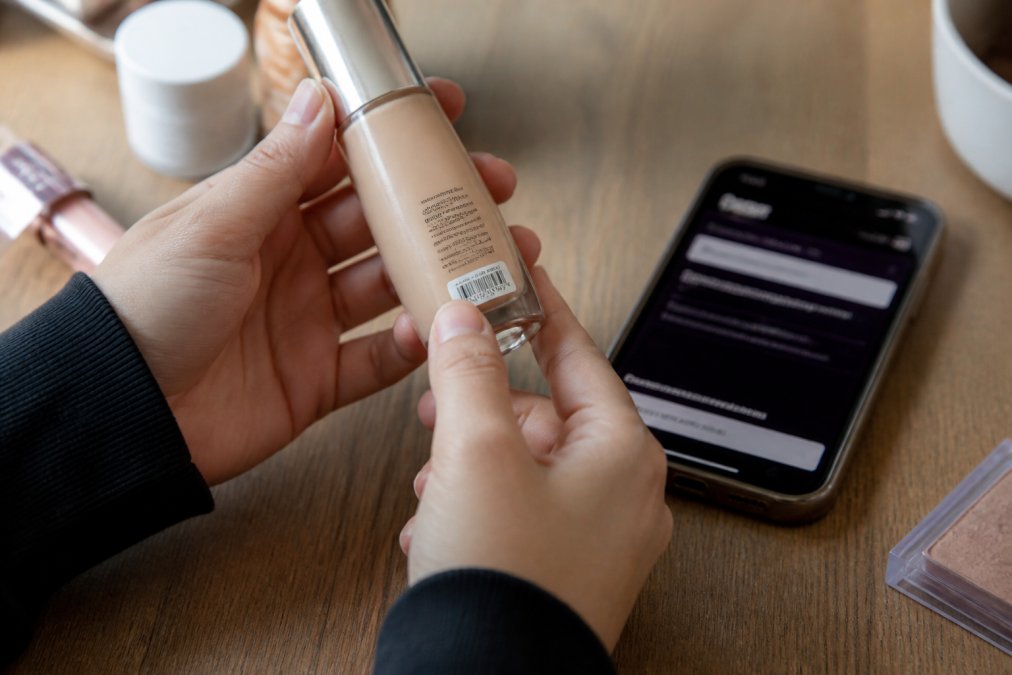 Close-up of hands holding a beauty product bottle, examining the batch code label under natural light, with a smartphone displaying a batch code verification website in the background