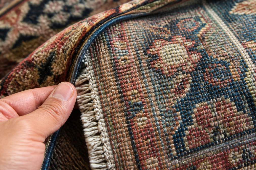 Close-up photograph of a hand-knotted rug showing the knot density, fiber texture, and intricate weaving pattern with natural lighting highlighting the craftsmanship and construction details