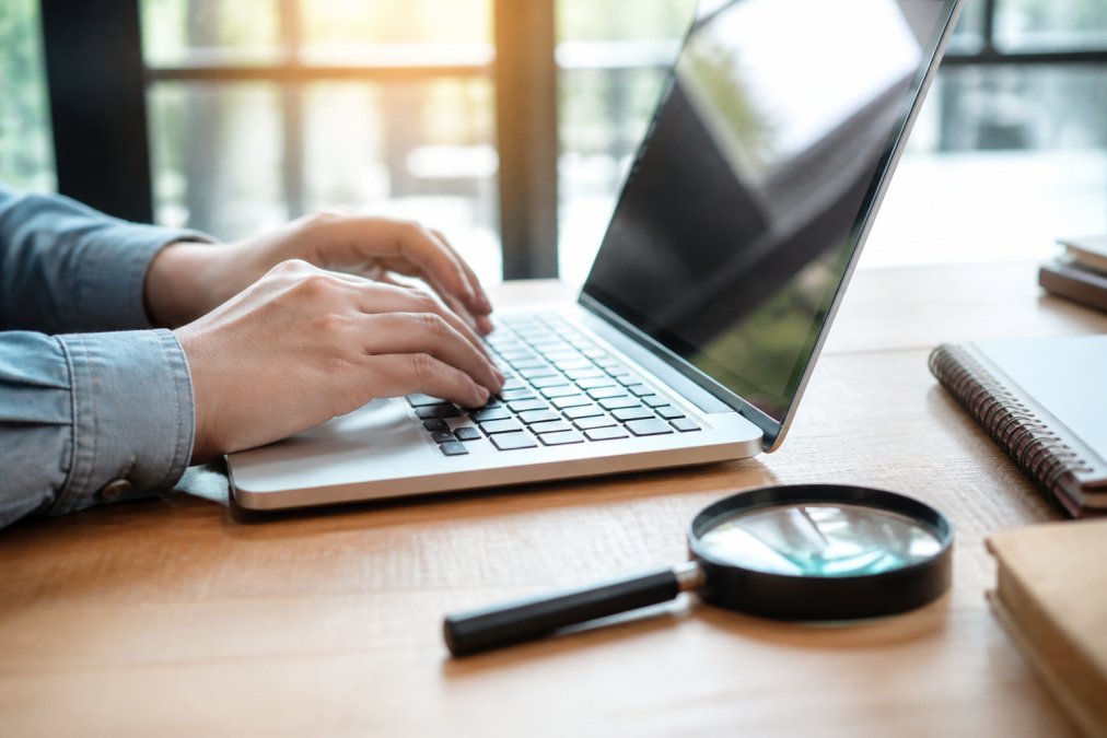 A close-up of a person's hands typing a website URL into a laptop browser, with a magnifying glass resting on the desk nearby, warm natural light from a window