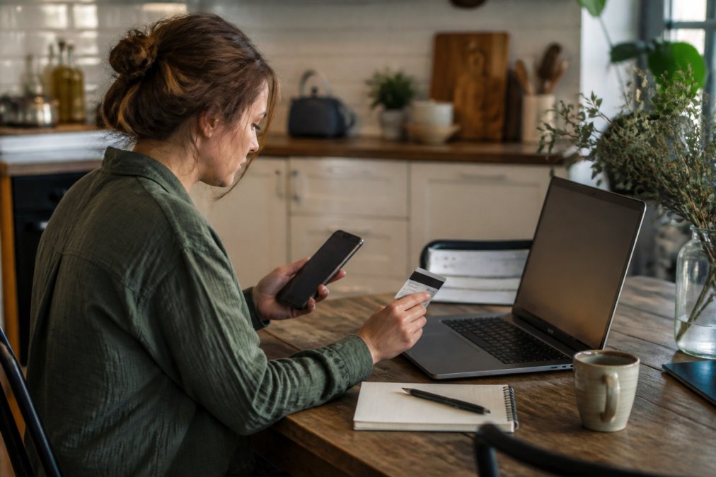 A person sitting at a kitchen table with a laptop open to a product review page, holding a smartphone in one hand and a credit card in the other, with a notepad and pen nearby for jotting down notes