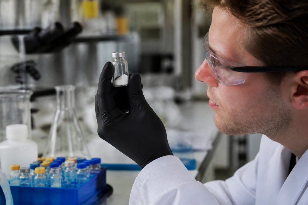 Close-up of a researcher in a white lab coat examining a small glass vial containing white lyophilized powder under bright laboratory lighting, with other vials and lab equipment visible in the background