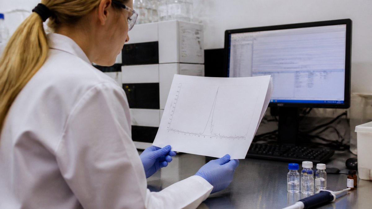 A researcher in a lab coat examining a printed HPLC chromatogram next to a computer screen showing analytical software, with glass vials and a pipette on the stainless steel bench under bright lab lighting