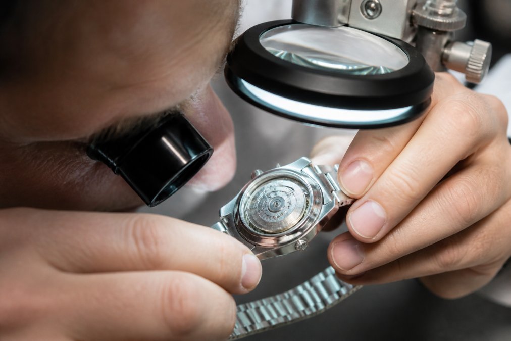 Jeweller using a loupe to examine detailed engraving on a luxury watch caseback under bright magnification lighting, showing precise inspection of serial number and hallmarks
