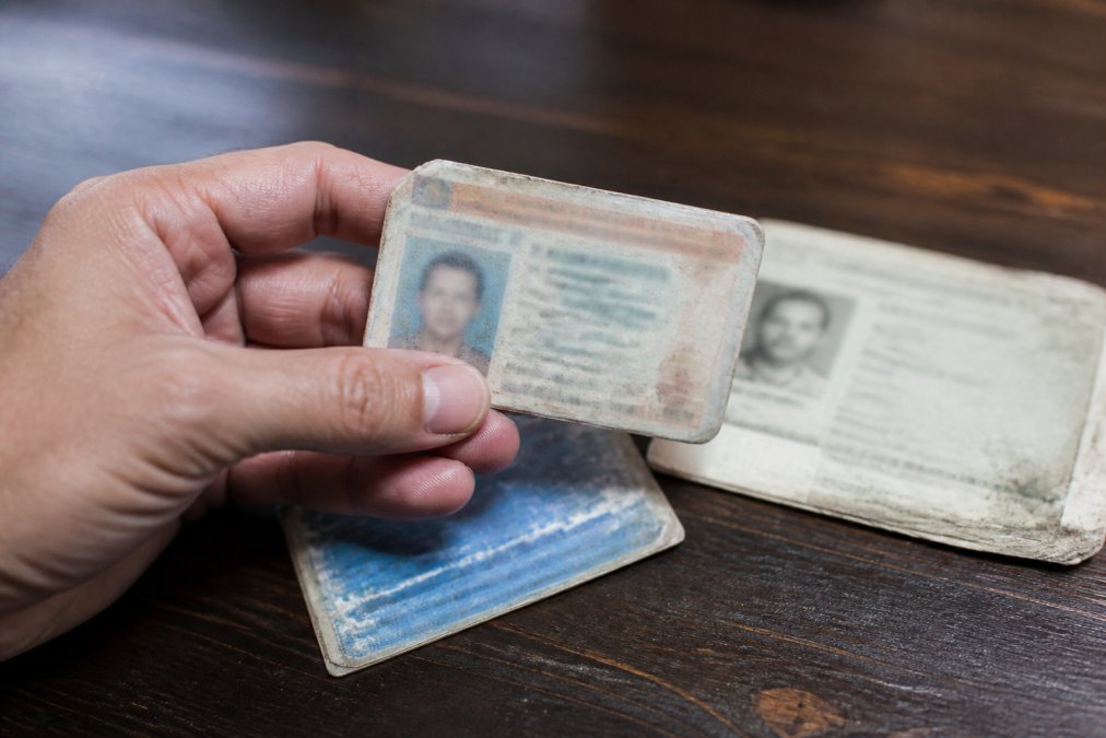 Close-up of a hand holding an expired driver's license and faded credential documents on a dark wooden table with harsh overhead lighting