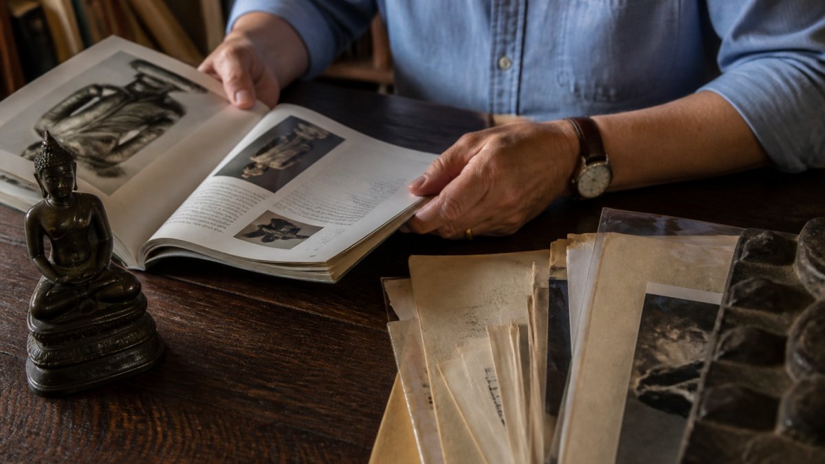 Les mains d'un collectionneur examinent un ancien catalogue de vente aux enchères et un dossier de documents de provenance à côté d'une petite statue bouddhiste en bronze sur un bureau en bois, dans une lumière naturelle et chaleureuse