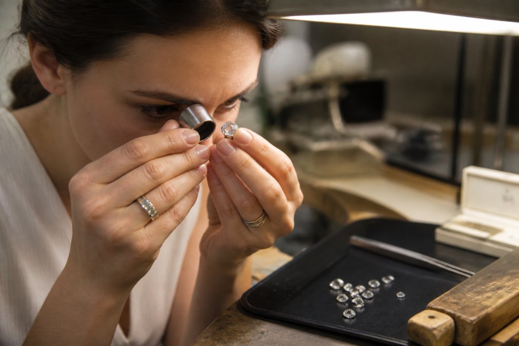 Professional jeweler using a jeweler's loupe to examine a moissanite stone under bright task lighting, with the loupe magnifying facet edges and light refraction patterns clearly visible