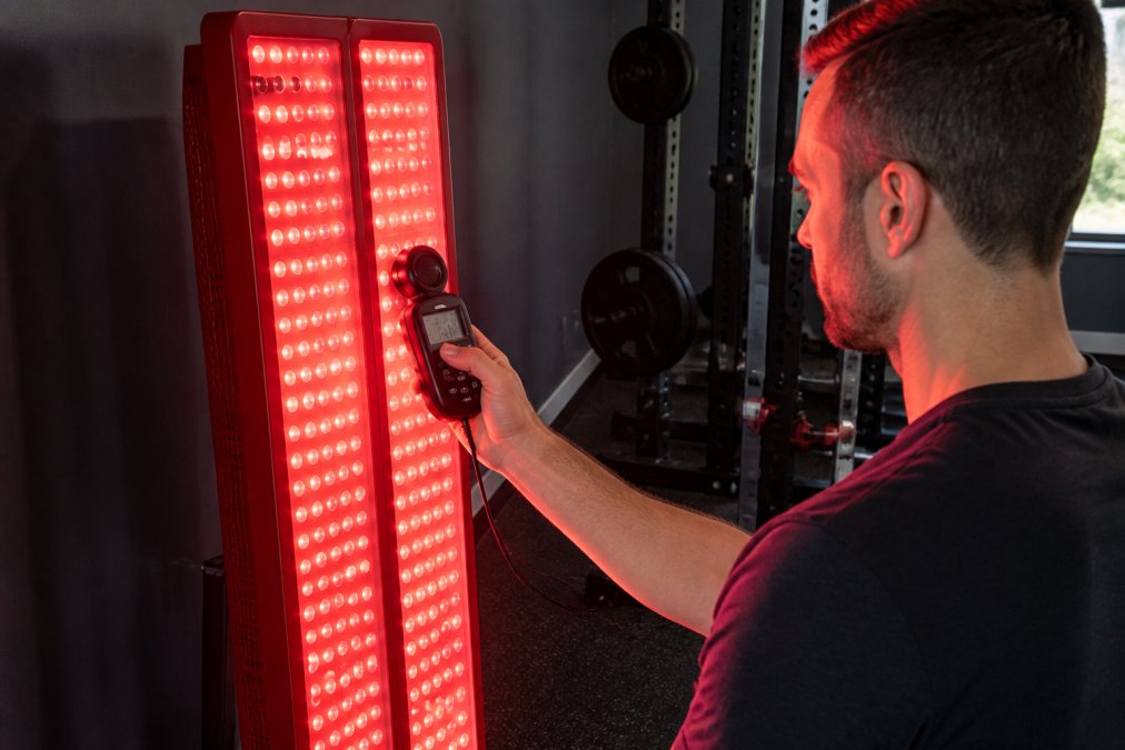 A person holding a small handheld spectrometer up to a large red light therapy panel in a home gym, the panel glowing bright red against a dark wall