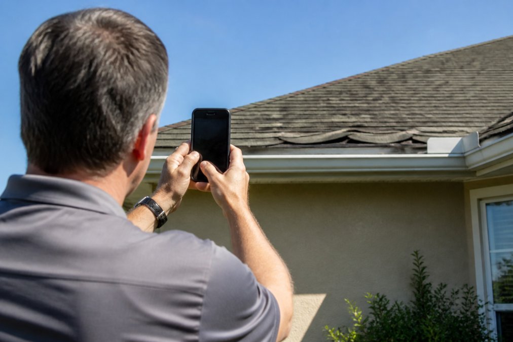 Close-up of homeowner photographing roof damage with a smartphone while standing safely on the ground near the property