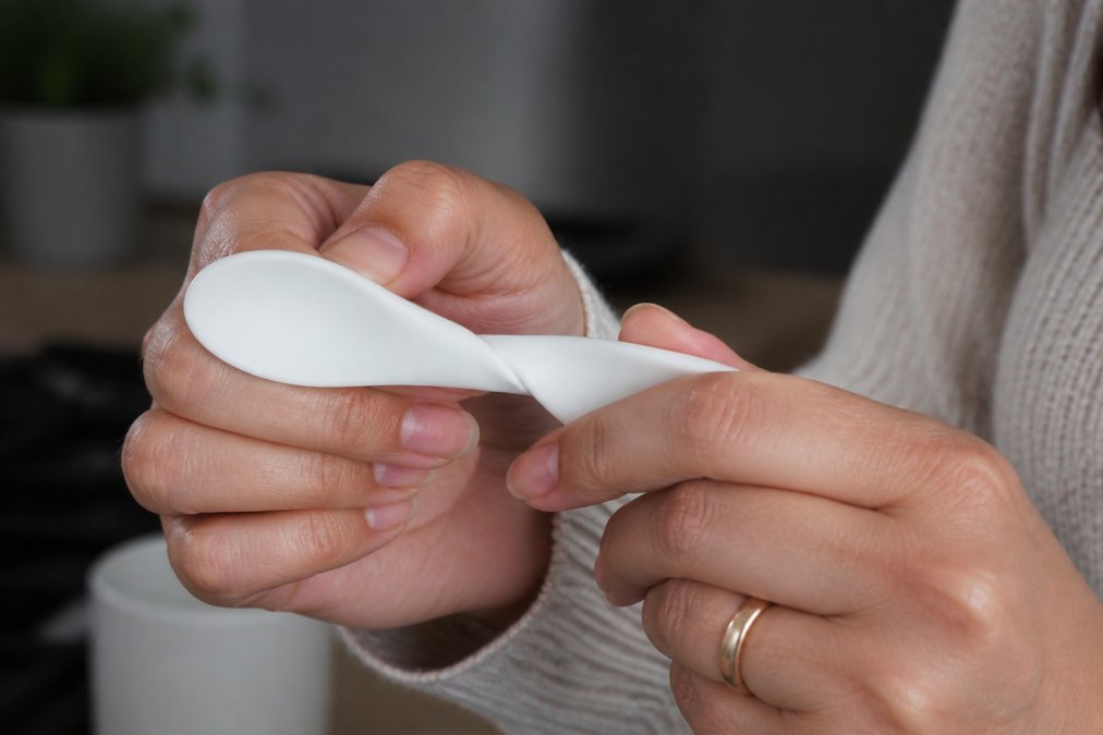 Close-up of a parent's hands performing the pinch and twist test on a white silicone feeding spoon, the material twisted and showing an opaque white colour change at the pinch point