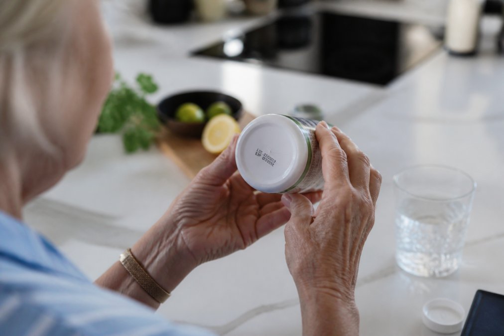 Close-up of an older woman's hands turning a white supplement bottle to read the small printed batch number and best-before date on the base label, on a kitchen worktop in natural daylight