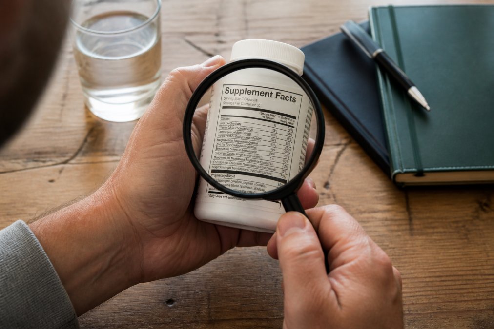 Close-up of a person's hands holding a supplement bottle, examining the Supplement Facts panel with a magnifying glass on a wooden table