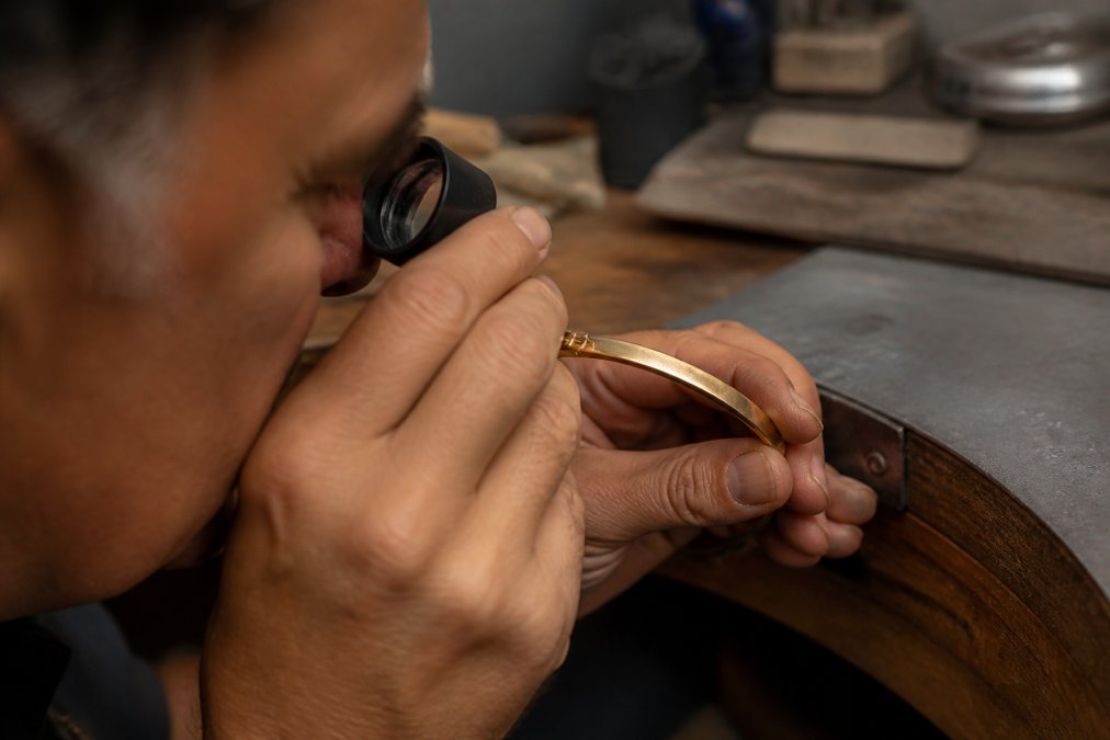 Jeweler using a 10x magnification loupe to closely examine hallmarks and maker's marks stamped inside a gold bracelet under bright, focused light on a jeweler's work surface