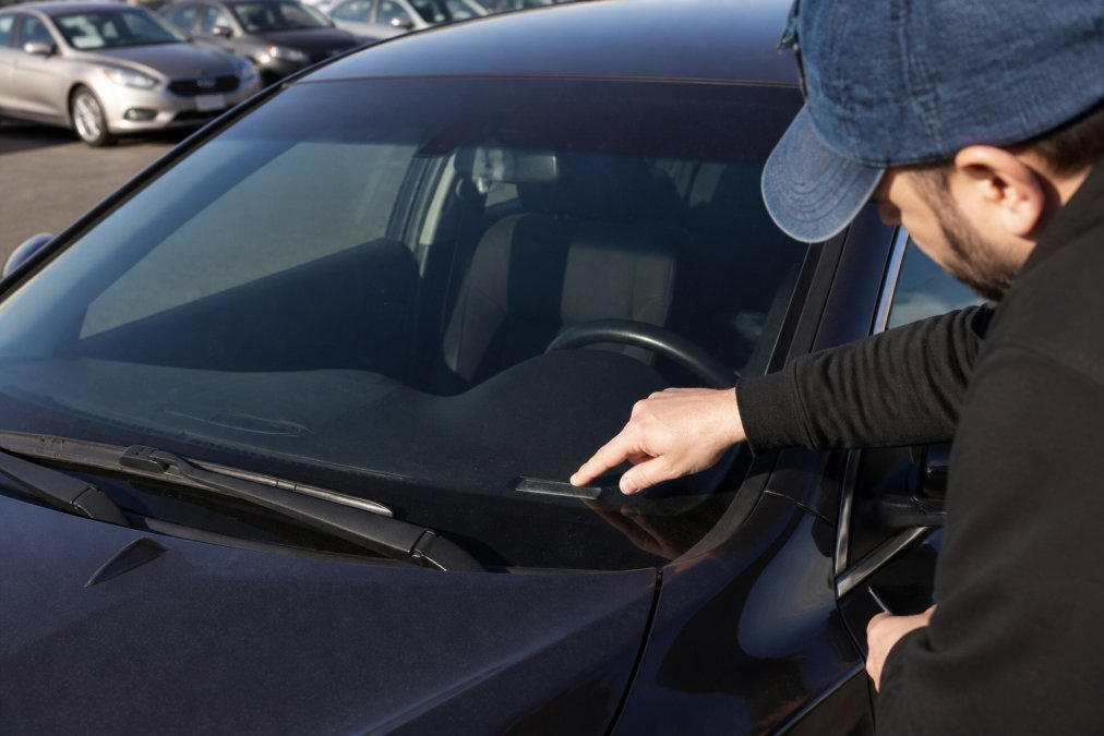 Close-up of a buyer's hand tracing the VIN plate on a used sedan's dashboard through the windshield at a dealership lot, afternoon light