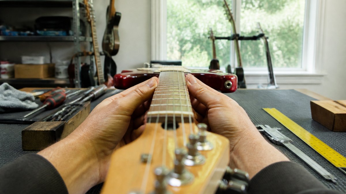 Close-up of hands holding a guitar at eye level while sighting down the neck from headstock to body, checking for straightness against natural light