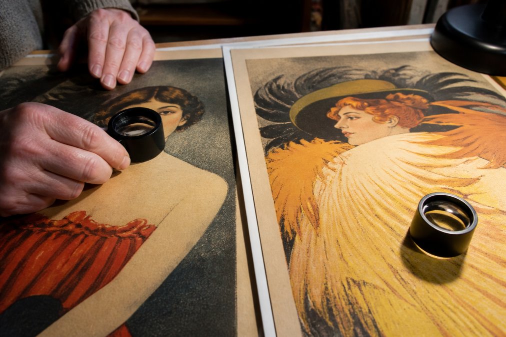 Close-up macro photograph of a collector's hands holding a magnifying loupe over a vintage poster surface, examining ink texture under warm desk lamp lighting