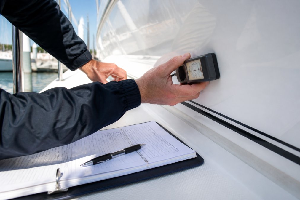 A marine surveyor's hands holding a moisture meter against a white fiberglass hull, with an open survey document and pen resting on the deck nearby in bright daylight