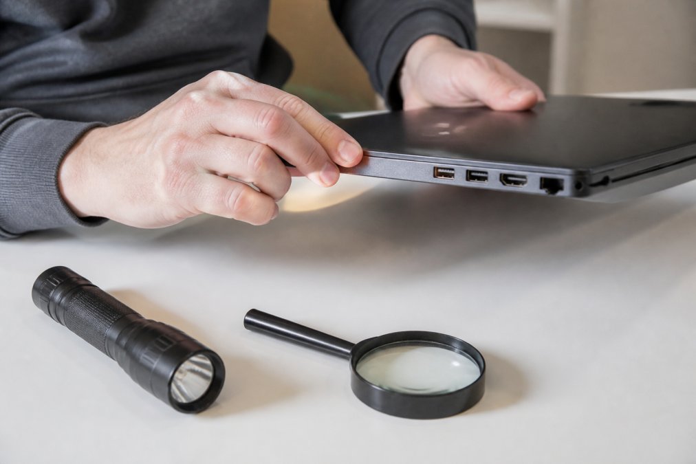 Close-up of hands inspecting a laptop's ports and casing for scratches or dents, with a flashlight and magnifying glass nearby on a clean desk