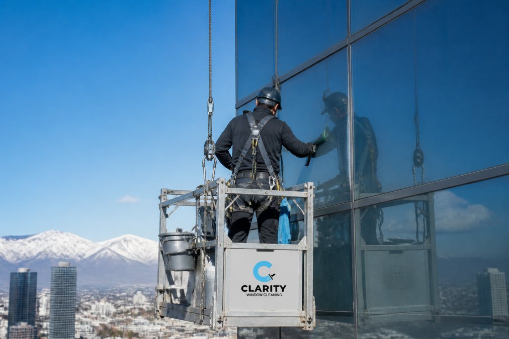 Professional window cleaner wearing full safety harness and fall protection equipment suspended on stage scaffold against modern high-rise glass facade with clear blue sky