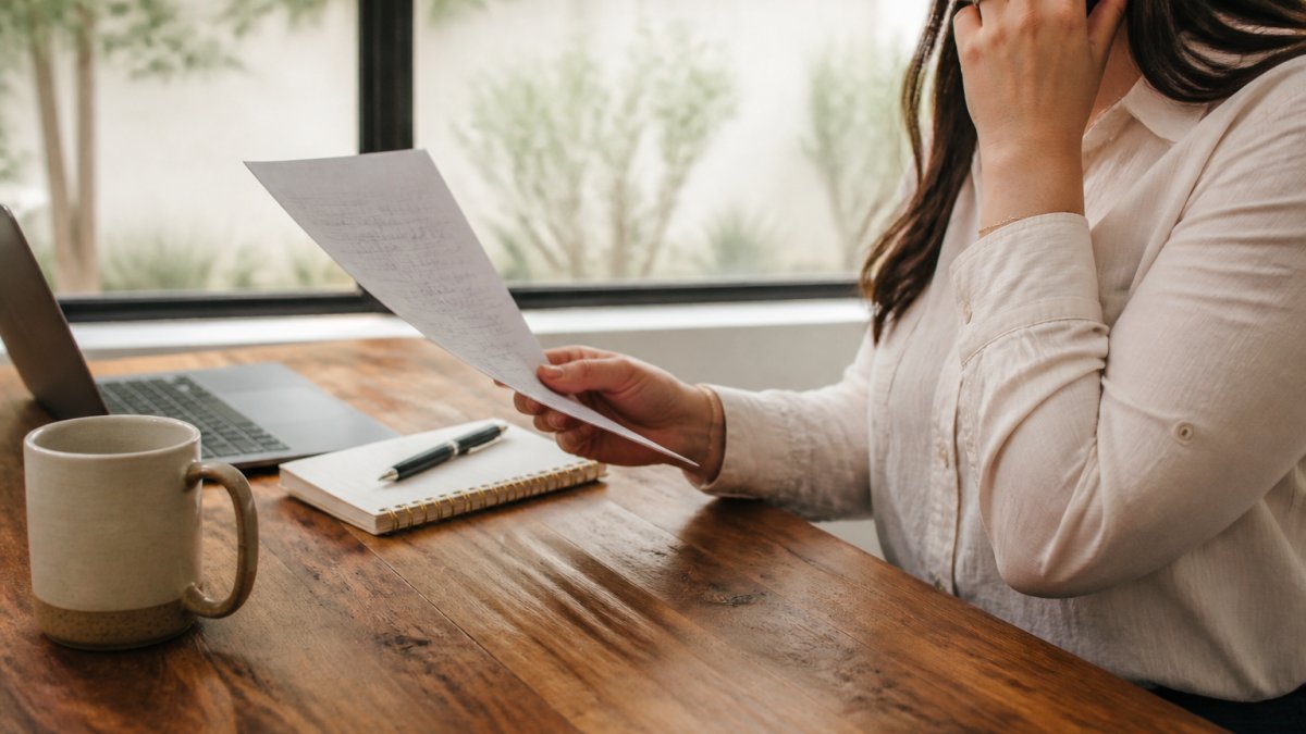 A person sitting at a desk with a laptop, notebook, and coffee, reviewing a handwritten list of questions on paper while talking on a phone call, warm afternoon light through a window
