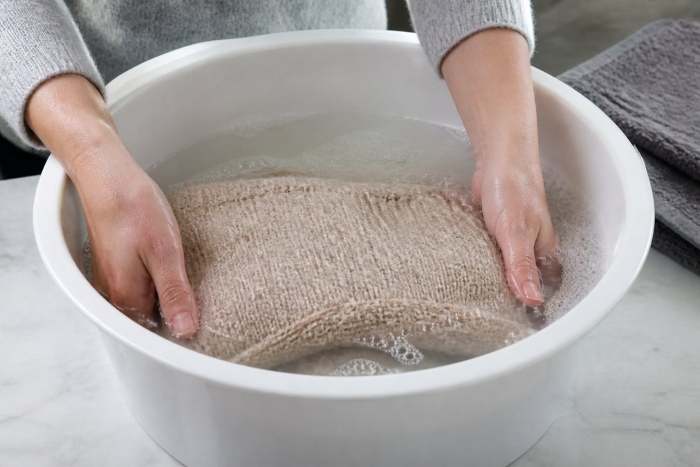 Hands gently immersing a folded alpaca sweater in a basin of cool water with mild detergent, showing proper submersion technique without wringing