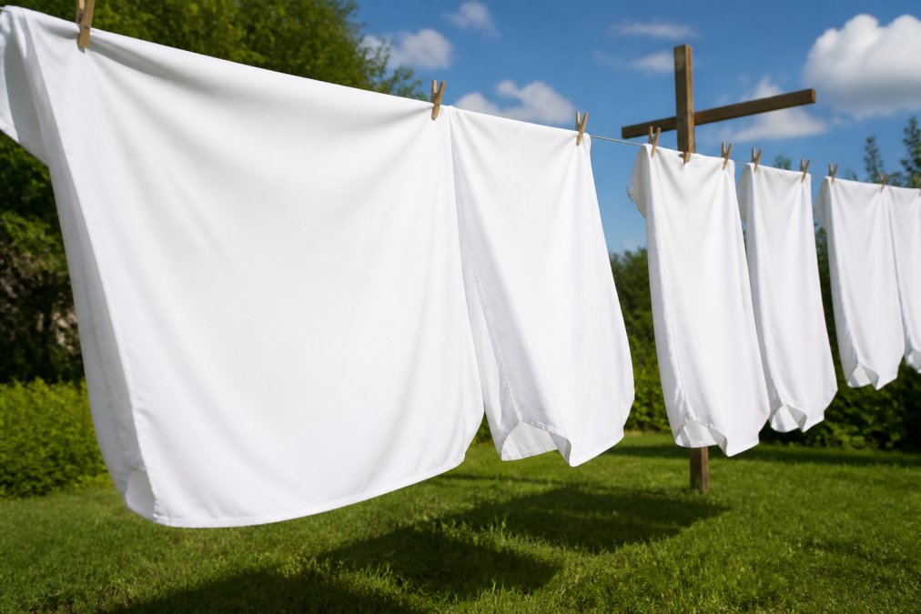 Freshly washed white bamboo viscose sheets hanging on an outdoor wooden clothesline in bright natural sunlight, gently billowing in a light breeze against a clear blue sky, green grass visible below