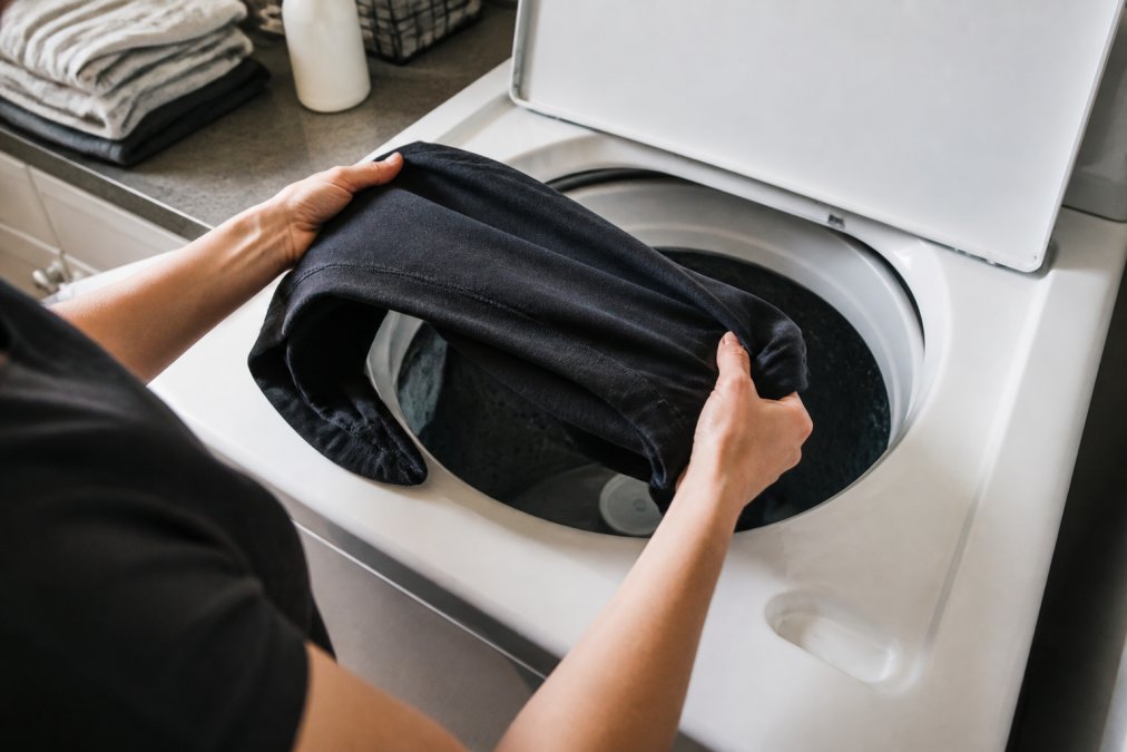 Close-up of hands turning a graphic sweatshirt inside out before placing it into a top-loading washing machine, bright laundry room lighting