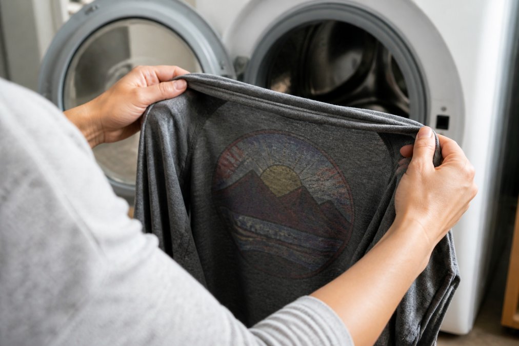 Close-up of hands demonstrating turning a graphic tee inside out to reveal the printed design on the inside, with a washing machine visible in the soft-focus background