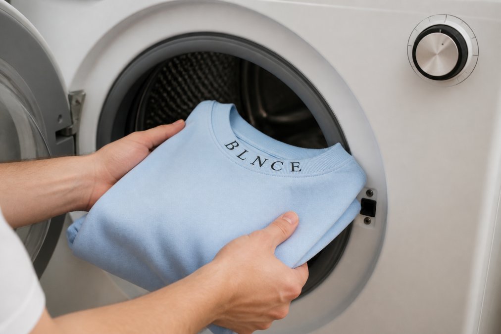 Close-up of hands loading heavyweight cotton garments into a washing machine, showing proper garment placement with the machine dial set to delicate cycle with natural lighting