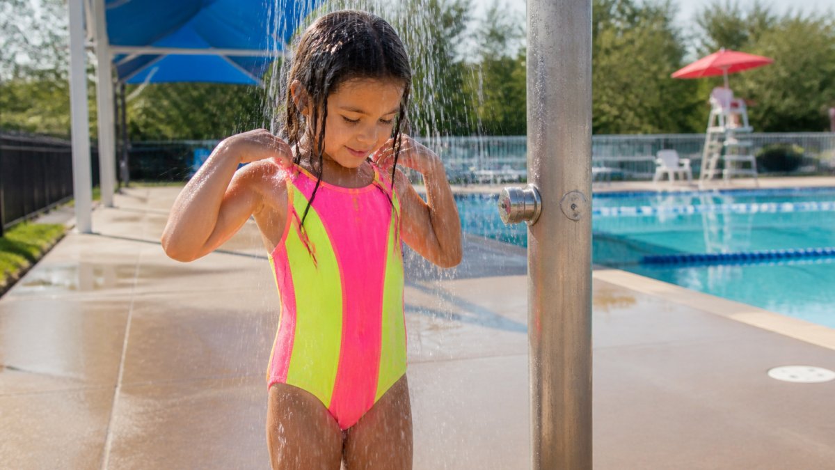 Child in bright neon swimsuit standing under an outdoor rinse station at a pool deck, fresh water streaming over the fabric, morning sunlight illuminating the vivid colors