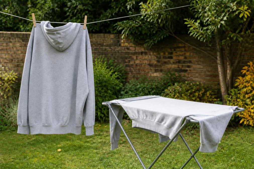 An oversized hoodie hanging on a clothesline in natural daylight, with a second hoodie draped flat on a drying rack nearby, both in the process of air drying
