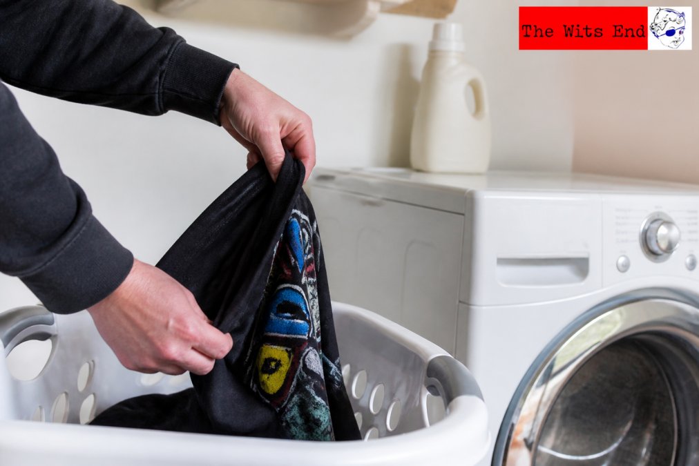 Close-up of a pair of hands turning a colorful graphic tee inside out over a white laundry basket, with a bottle of mild liquid detergent sitting on top of a front-load washing machine in a bright laundry room