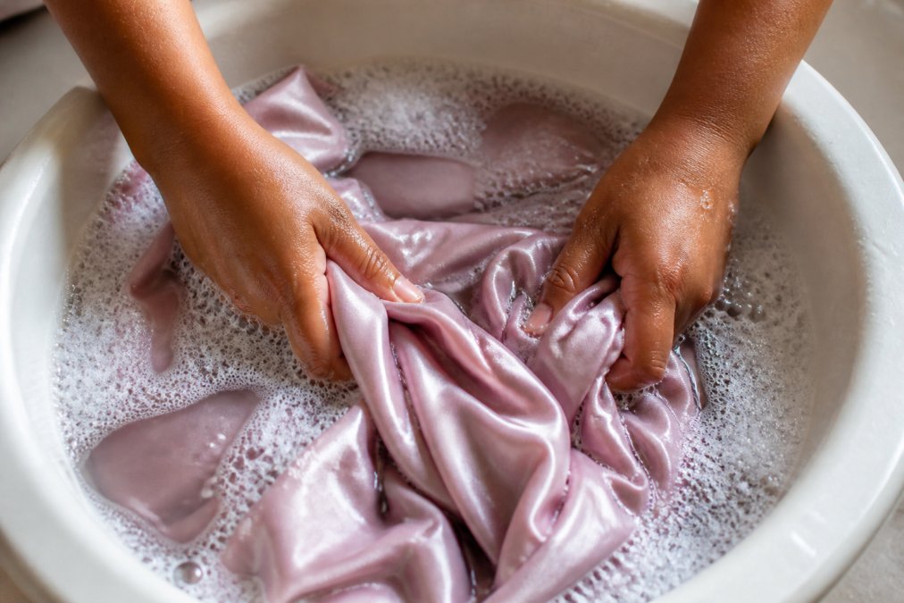 Close-up of hands gently agitating a luxurious silk robe in a basin of tepid water with soap suds, showing proper delicate hand washing technique in natural light