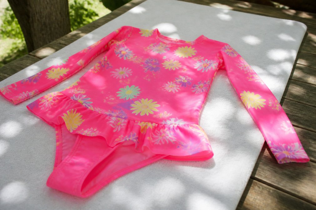 Bright-colored UPF swimsuit laid flat on a white towel in shaded outdoor area, with dappled sunlight filtering through tree leaves in background, no direct sun exposure