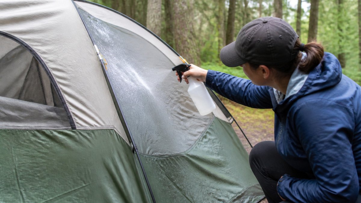 Person spraying water on tent exterior with spray bottle during daylight to test for leaks, examining fabric and seams for water intrusion