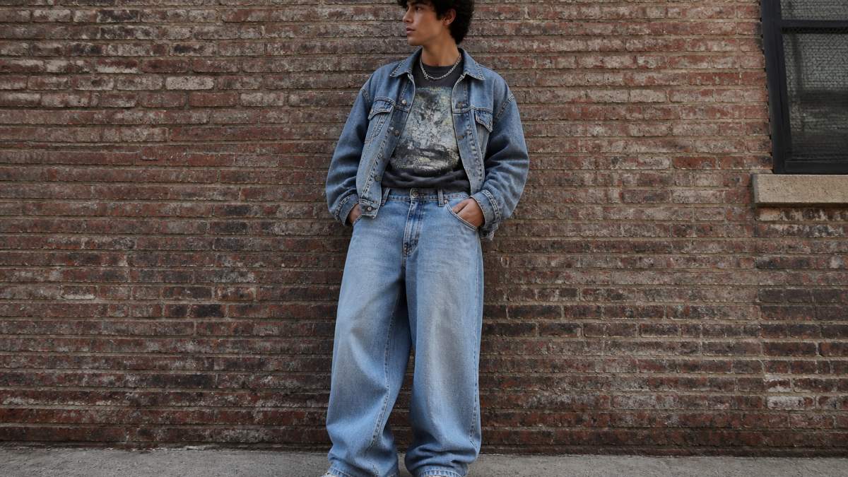 Young person wearing oversized washed graphic tee tucked into baggy denim, layered with a denim jacket, wearing white chunky trainers, standing in natural daylight against a neutral urban brick wall