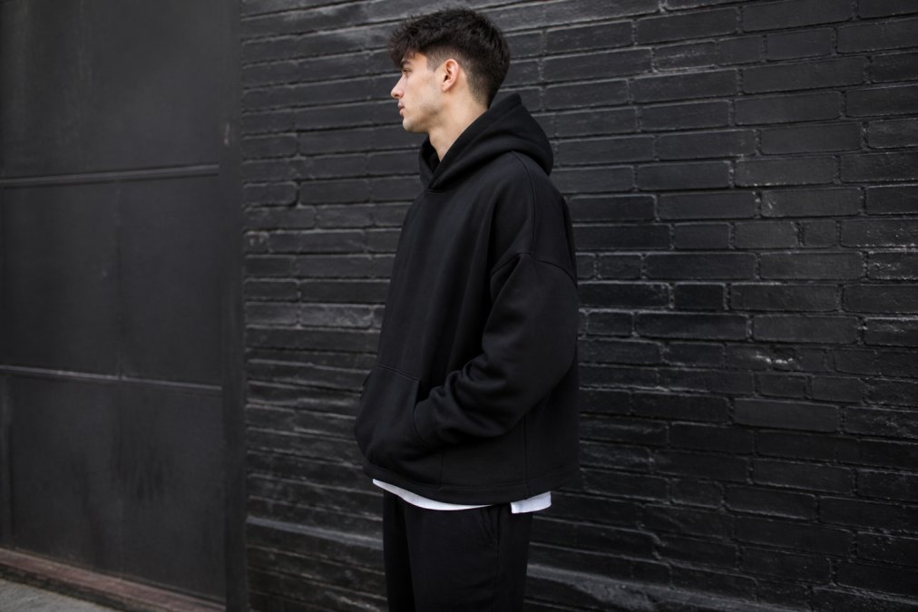 A young man wearing an oversized black hoodie with drop shoulders, standing against an urban brick wall, showing the boxy fit and relaxed silhouette from the side, natural daylight