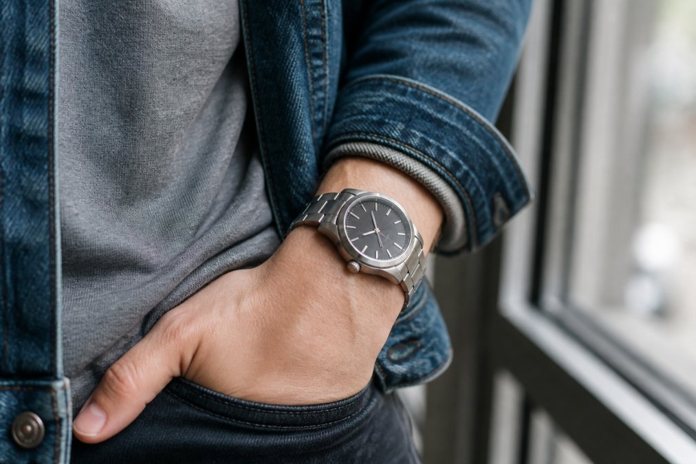 Close-up of a wrist wearing a titanium watch with a brushed finish, paired with a denim jacket sleeve and a simple grey t-shirt, shot in soft natural window light
