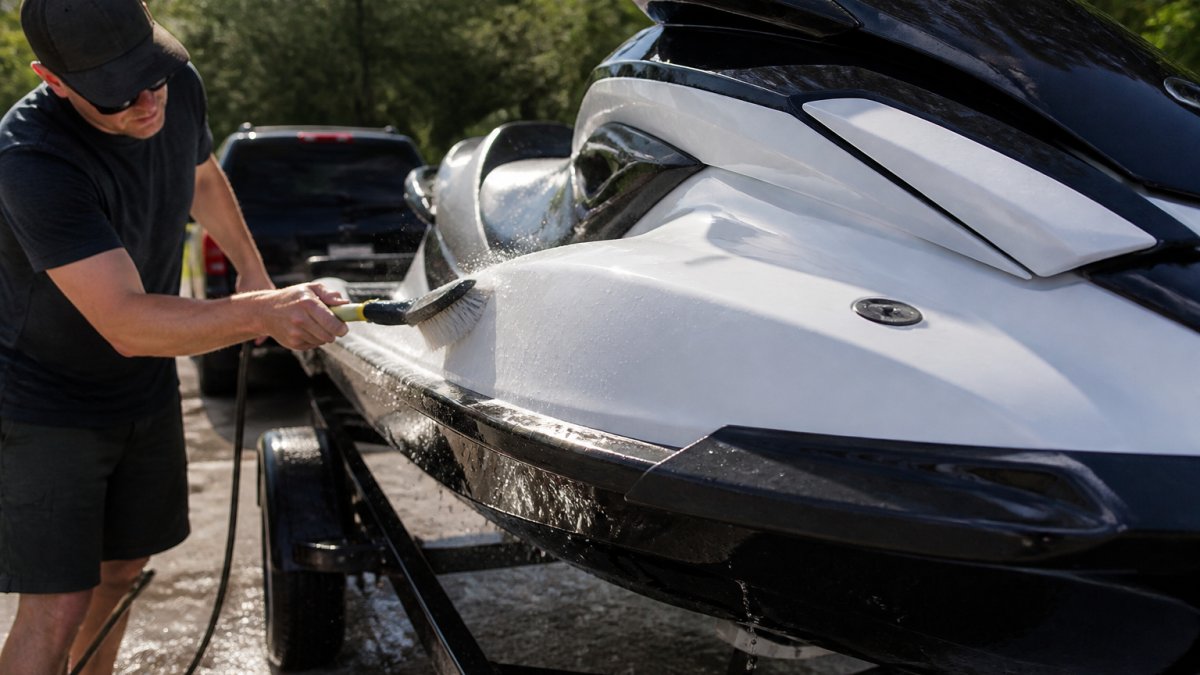Person cleaning the exterior of a jet ski hull with a soft brush under natural sunlight, with water spraying across the white fiberglass surface