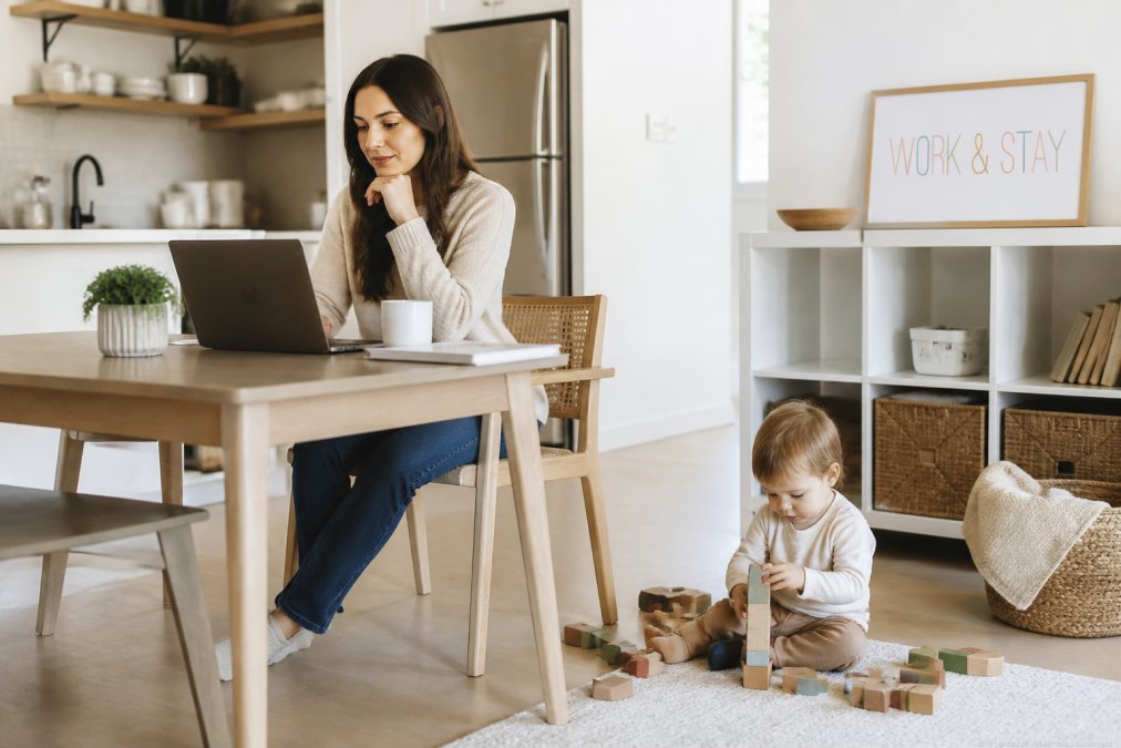 A parent working on a laptop at a kitchen table while a toddler plays with blocks on the floor nearby, natural daylight, organized home workspace
