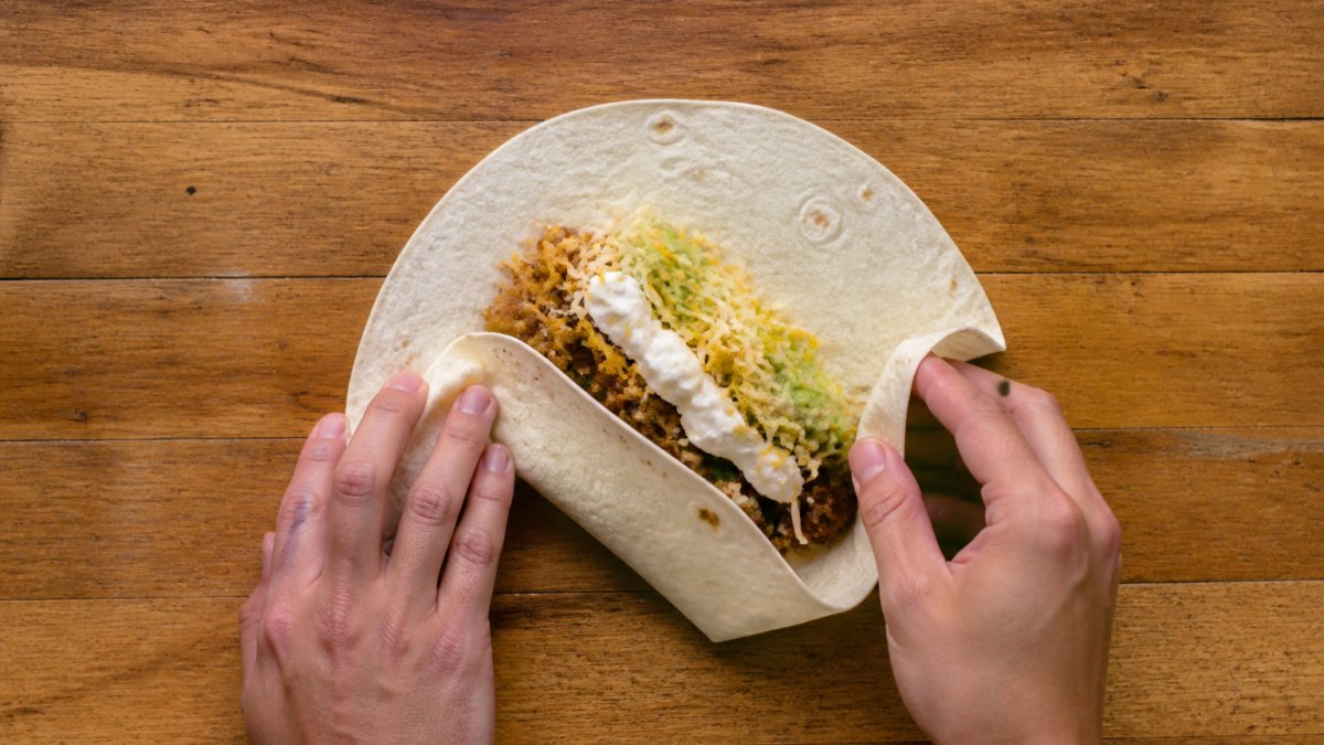 Hands demonstrating the step-by-step process to wrap a burrito on a wooden surface with fresh ingredients.