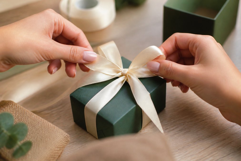 Close-up of hands demonstrating ribbon bow technique on a small jewelry box, showing the ribbon loops being formed and the final knot detail with precise finger positioning