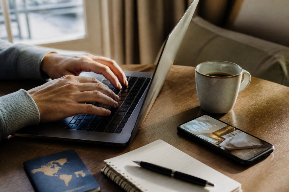A close-up of hands typing an email on a laptop with a hotel booking confirmation visible on a smartphone screen nearby, warm natural light from a window, coffee cup on the desk
