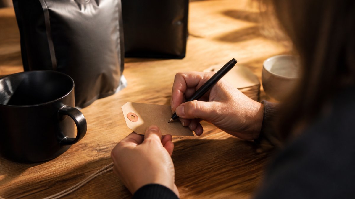 Close-up of hands writing a short message on a kraft paper coffee gift tag with a fine-tip pen, a freshly roasted coffee bag blurred in the background, warm natural light