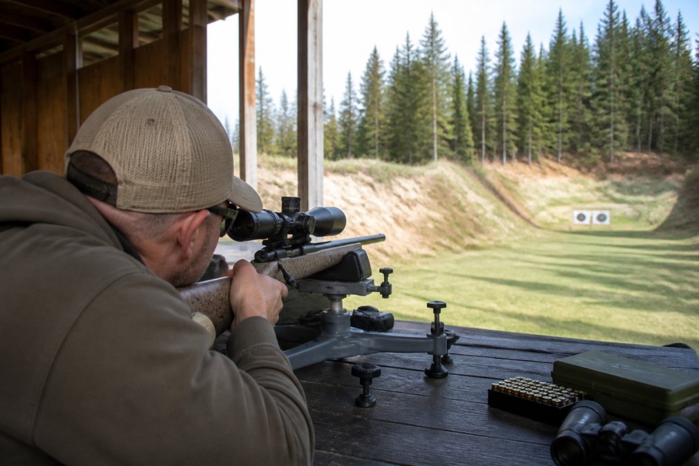 Shooter positioned at bench rest with rifle mounted in shooting rest, aiming downrange at target 200 yards away, scope turrets visible, range equipment and targets in background