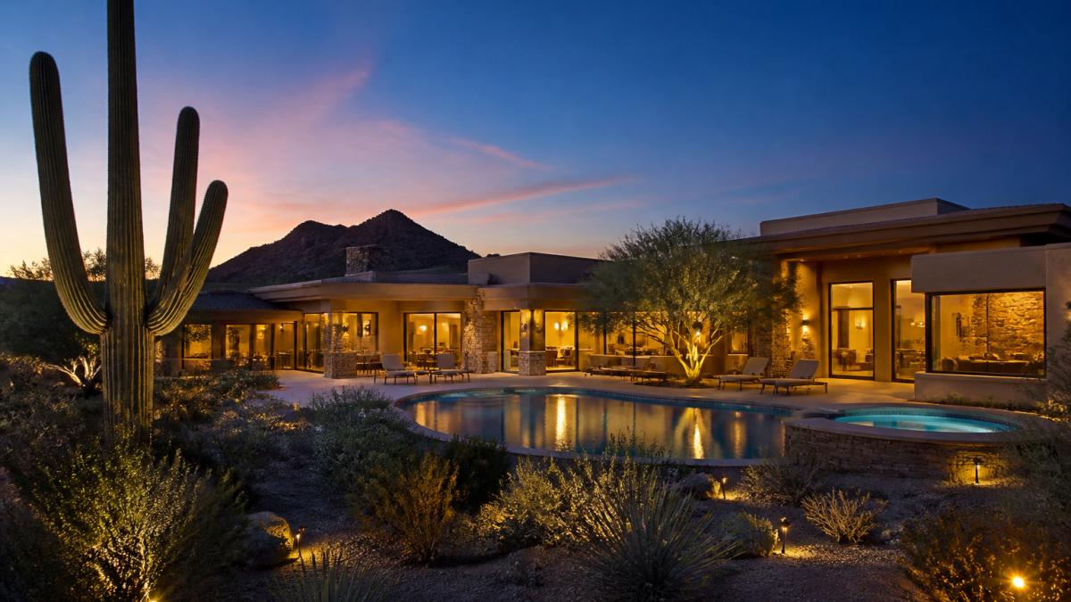 A luxury desert home exterior at twilight with warm interior lights glowing through large windows, a lit pool reflecting the deep blue sky, and saguaro silhouettes in the foreground