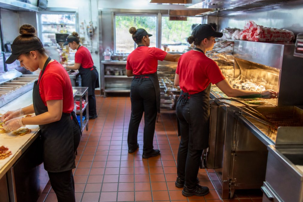 Interior view of a Wendy's kitchen during peak service, with multiple staff members managing food preparation stations and order fulfillment, natural lighting from service windows