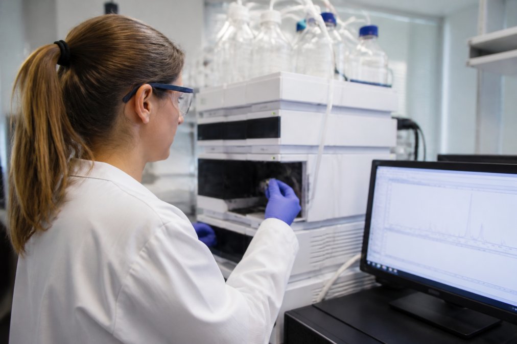 Laboratoriummedewerker die naar een HPLC-apparaat kijkt met een computermonitor met chromatogramgegevens en piekanalyse op de achtergrond, onder fel laboratoriumlicht